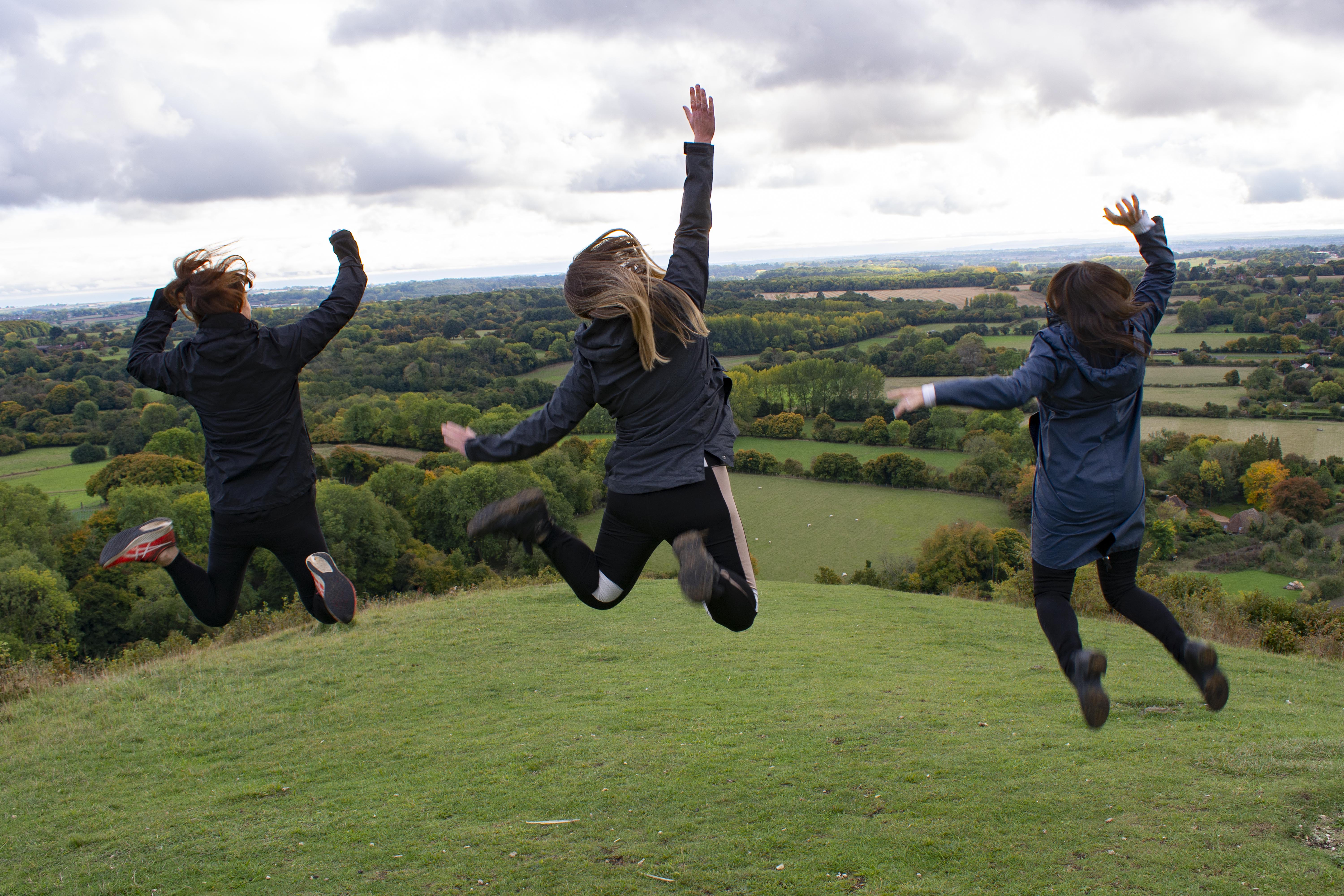 Three people jumping in the air, backs facing the camera as they smile, withA person looking out across rolling fields and scenery in the background.