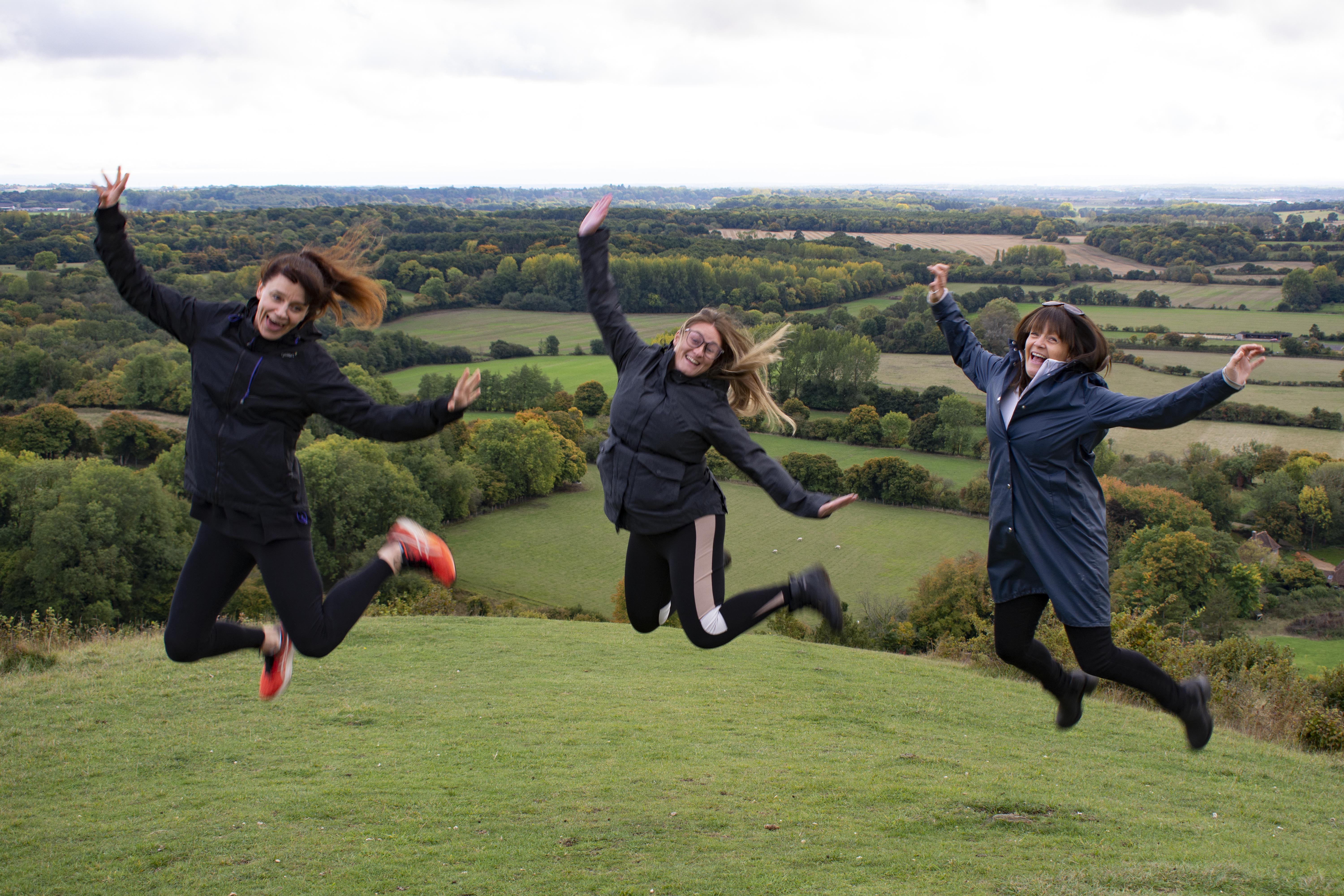 Three people jumping in the air, faces looking at the camera as they smile, withA person looking out across rolling fields and scenery in the background.