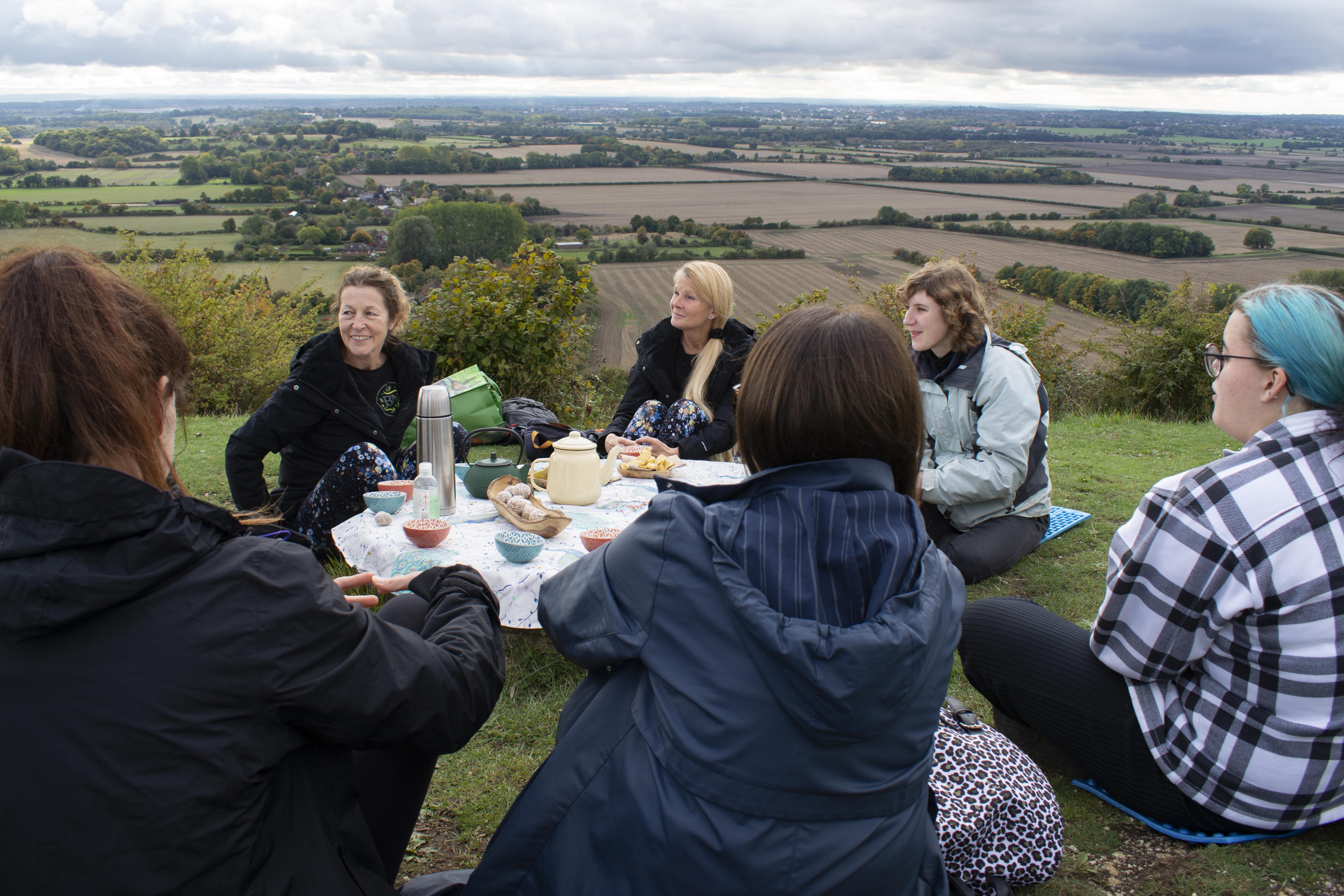 People sat around a treestump with a table cloth, a kettle and other amenities on it. Rolling fields and the sea can be seen in the background.